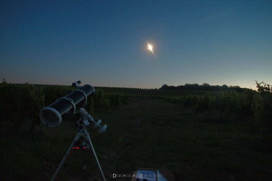 Paysage valloné avec des vignes au crépuscule, la Lune (surexposée) est visible juste au dessus du l'horizon. Télescope de couleur noir sur une monture équatoriale, à gauche.
