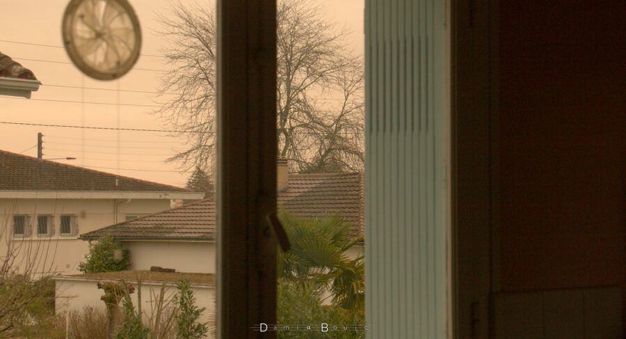 Vue resserrée au travers d'une fenêtre, montrant quelques maisons pavillonnaires, un arbre décharné et un ciel ocre
