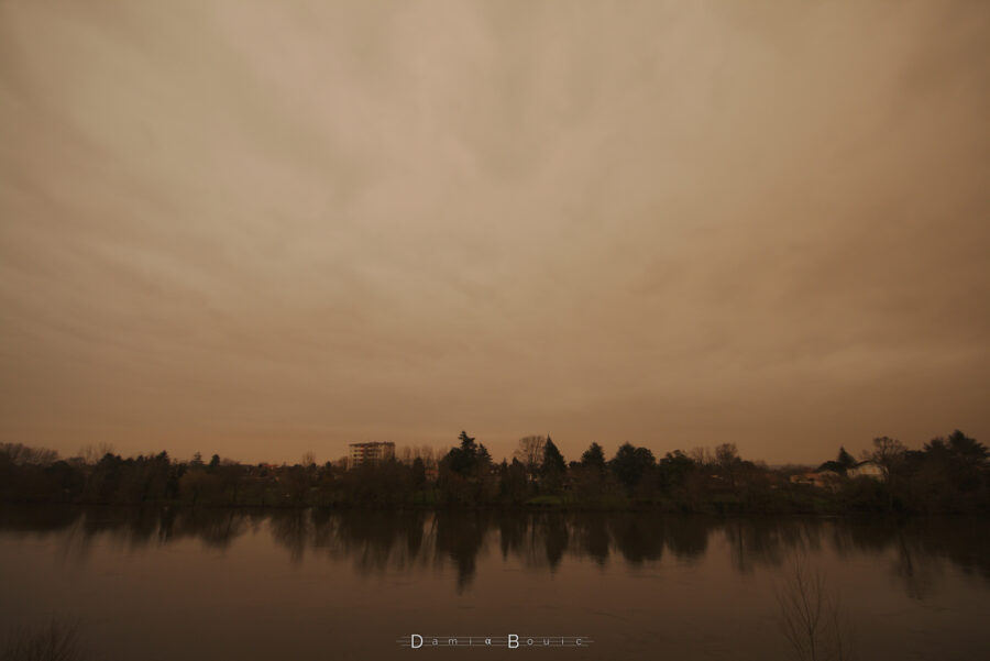 Photo en ultra-grand angle montrant le ciel jaune-orange, irrégulier, et du paysage en dessous (ligne d'horizons constituée majoritairement d'arbres, d'un seul immeuble de 6 étages, le tout se reflétant dans l'eau de la Dordogne)