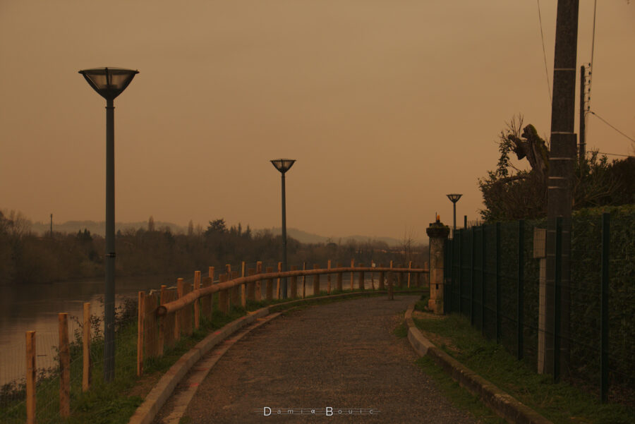 Nouveau zoom avec chemin en bitume défoncé, lampadaires, et paysage lointain, avec une haie à droite qui borde le tout