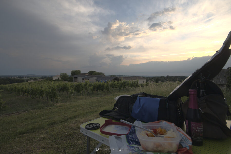 Premier plan, table de camping à droite avec un sac photo ouvert, un plexi ouvert avec une salade de tomates-mozza et une fourchette plongée dedans. Une bière aromatisée à la framboise est visible à droite. Couvercle et bouchon d'objectif trainent sur le coin gauche de la table. Paysage : herbe rase, vignes qui descendent, bâtiment viticole derrière. Horizon dégagé et ciel encombré, sombre à gauche plus clair à droite avec de nombreux morceau de nuages écorchés.