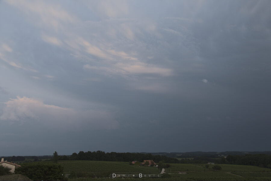 Image zoomée. Horizon semi-boisé. Des franges nuageuses saisissent les derniers rayons du Soleil. Le reste est uniformément gris.