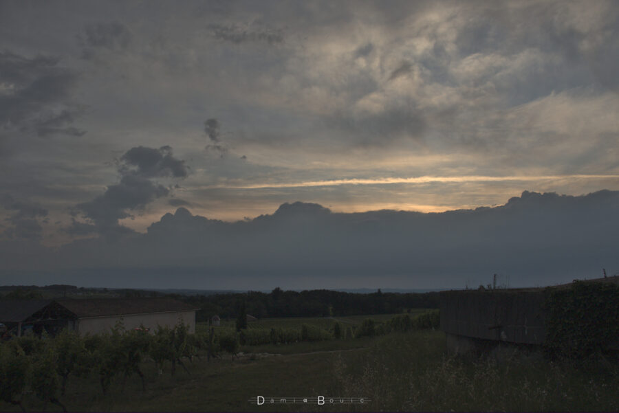 Le Soleil se couche mais il n'est pas visible, masqué par une barre de nuages sombre et compacte. Ces nuages sont boursoufflés par ailleurs. Le reste du ciel est teinté de lumière solaire et de rugosités nuageuses.