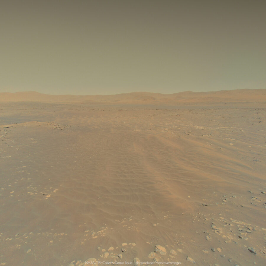 A picture from Ingenuity drone, showing the martian landscape from a few meters above the ground. We can see dunes, some boulders, and on the horizon, some hills