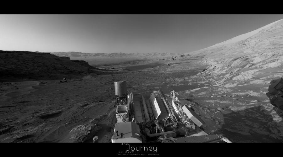 Panorama martien en niveaux de gris, avec une portion de ciel au premier tiers, le reste étant du paysage très contrasté du fait du Soleil bas sur l'horizon. A gauche une butte rocheuse aplatie projète son ombre sur le sol martien, qui est rocailleux et rugueux. Au centre, la vue porte sur une large plaine, un peu gondolée et présentant des dunes de sables bien visibles. A droite, l'horizon remonte brusquement au gré du flanc du Mont Sharp. Au milieu en bas, la partie arrière du corps du rover, avec son antenne en forme de cylindre, des panneaux de métal indiquant l'emplacement de la source électrique