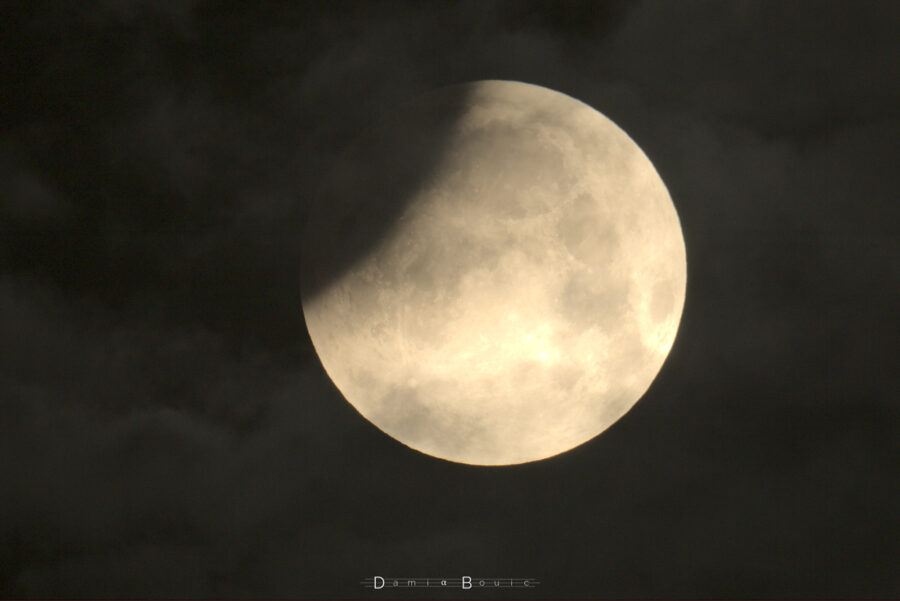 Disque lunaire dont la partie en haut à gauche est mordue par une ombre. quelques nuages semblent survoler la scène