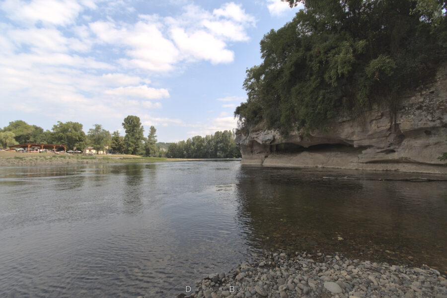 Quelques galets en bas du cliché s'enfoncent dans un eau translucide. Pas très loin, une petite falaise calcaire, dont le centre est entaillé par ce qui semble être une grotte, est surmontée d'arbres enchevetrés.