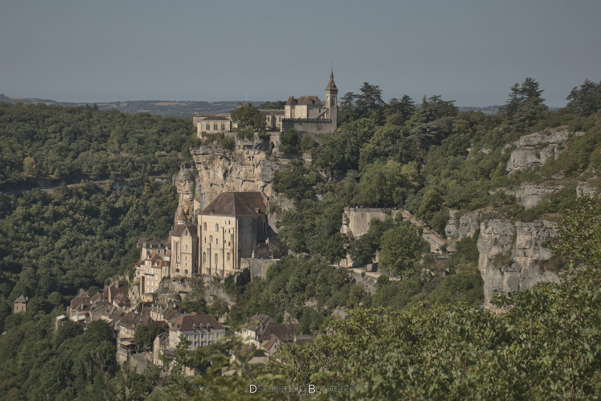 Cité médiévale accrochée à une falaise calcaire, un peu masquée par les arbres et les buissons. A gauche l'autre côté de la vallée, et au milieu l'horizon, lointain