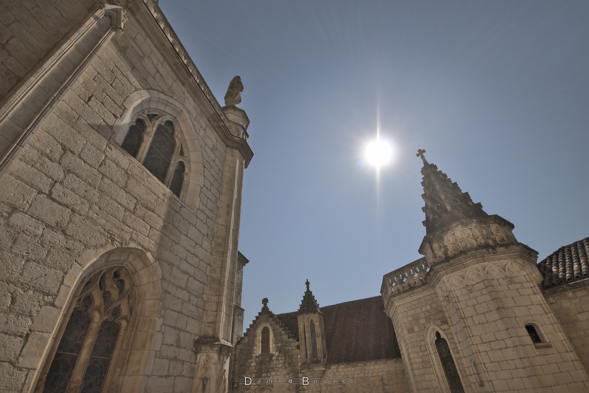 Contrejour avec le Soleil qui brille au centre. A gauche un pan de l'édifice religieux avec ses fenêtre sculptées et ses vitraux, à droite un petit clocher de pierre pyramidal, aux arrêtes dentelées