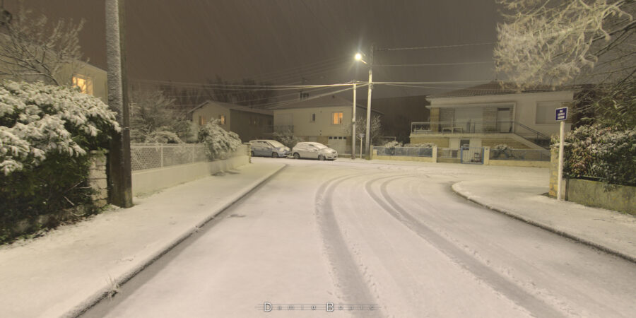 Vue d'une rue le soir, sous la lumière des lampadaires, avec les routes, les trottoirs, les toits, blanchis par une fine couche de neige.
