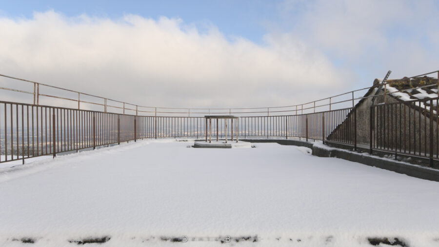 Environnement clos par de la cloture métallique, formant un arrondi. Au fond, une structure métalique sur laquelle repose une dalle en ciment. Le tout est enneigé, avec le sol lisse et d'un blanc éclatant.