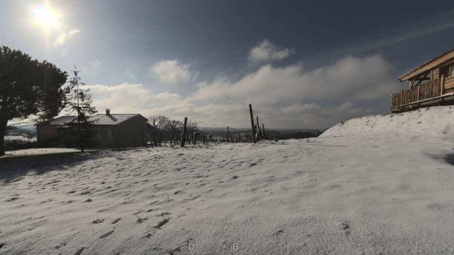 Neige au sol avec deux rangs de vigne qui émergent de la butte, une maison à gauche et uns des cabanons en bois sur la droite, plus haut.