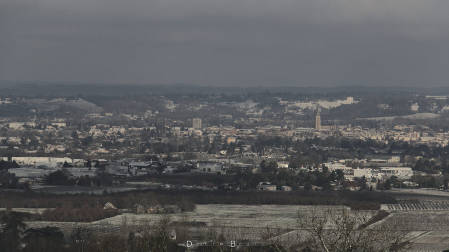 Photo fortement zoomée sur la ville de Bergerac, dont on distingue le clocher de l'église, comme une pointe vers le ciel, entourée de toits blanchis et d'autres batiments s'étirant de toutes parts. A l'arrière, les collines du nord de la ville, couvertes de forêts et dont les parties clairsemées sont blanches.