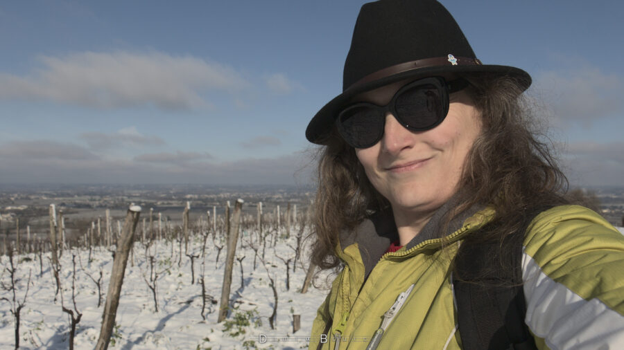 Photo de mon visage, très souriant, qui exprime la joie et la satisfaction de voir de la neige chez moi. Je porte un chapeau noir, avec un petit autocollant Tiplouf sur le cuir, je porte des lunettes noires. Mes longs cheveux ondulés blonds dépasses sur mes côtés, et je porte un manteau épais. Derrière moi, les vignes sous la neige.