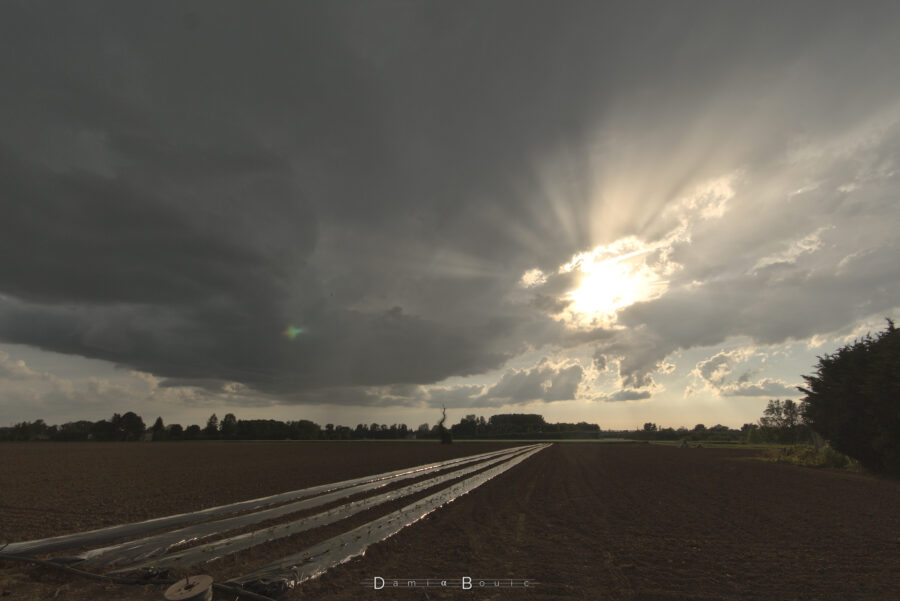 Vue plus large, les nuages envahissent tout le ciel, le Soleil perce au travers des nuages à droite, avec de nombreux rais de lumière rendus visibles par les premières pluies émises par l'orage situé plus à gauche. On voit dans le champs labouré les quatre bâches plastique noires qui filent vers l'horizon.