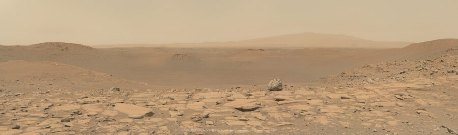 Panorama martien montrant un cratère peu profond parcouru de rides de sables, avec de douces collines dans le lointain. L'avant plan est parcouru de quelques rochers