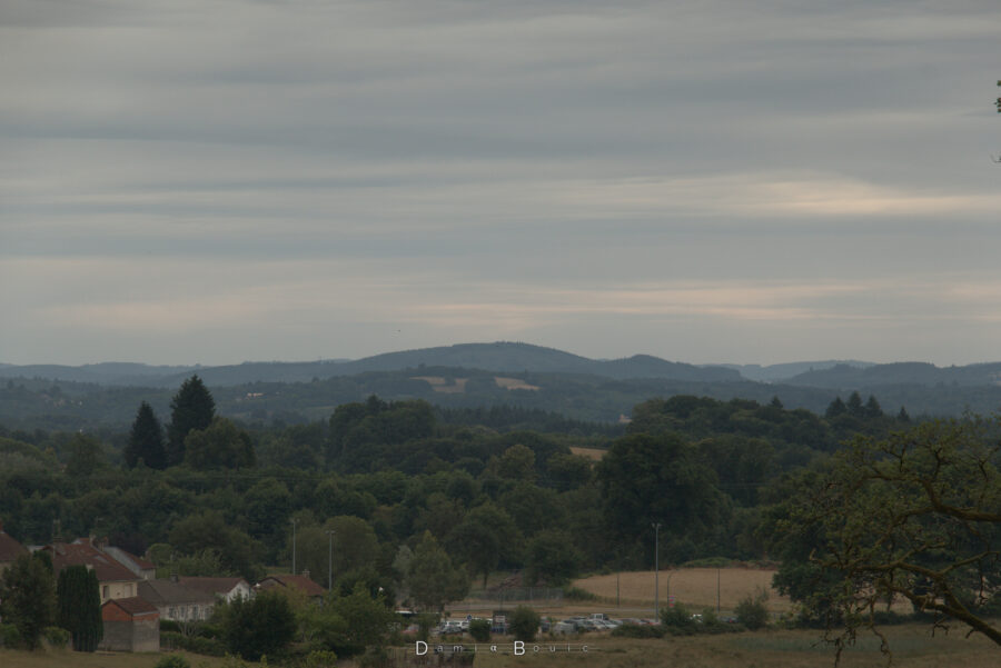 Vue sur un paysage boisé et quelques champs, la vue porte loin. Absorbées par la brume, plusieurs collines sont visibles sur l'horizon. Le ciel est couvert de nuages striés et ondulés, procurant une ambiance assez lourde.