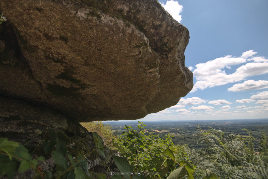 Photo prise sous la fameuse roche branlante, qui est assez plane en dessous, sur un large point de contact. Quelques fougère à la base et au fond le paysage lointain.