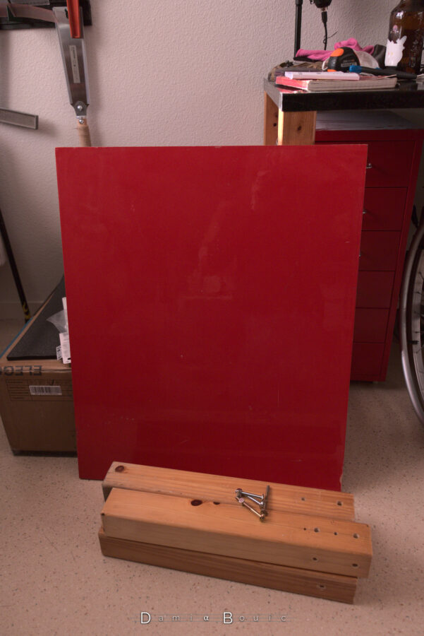 Une planche rouge rectangulaire, posée sur la tranche, contre le pied de mon bureau, avec au sol juste devant 4 poutrelles en bois de profil carré, et les vis qui serviront à l'assemblage dessus.