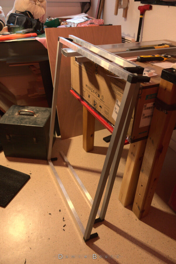 Les poutrelles sont assemblées partiellement, on voit deux cadres rectangulaires, dont les angles sont constitués de genre de chevrons en plastique noir. Les deux rectangles reposent l'un sur l'autre, à la verticale, posés contre le carton de l'imprimante