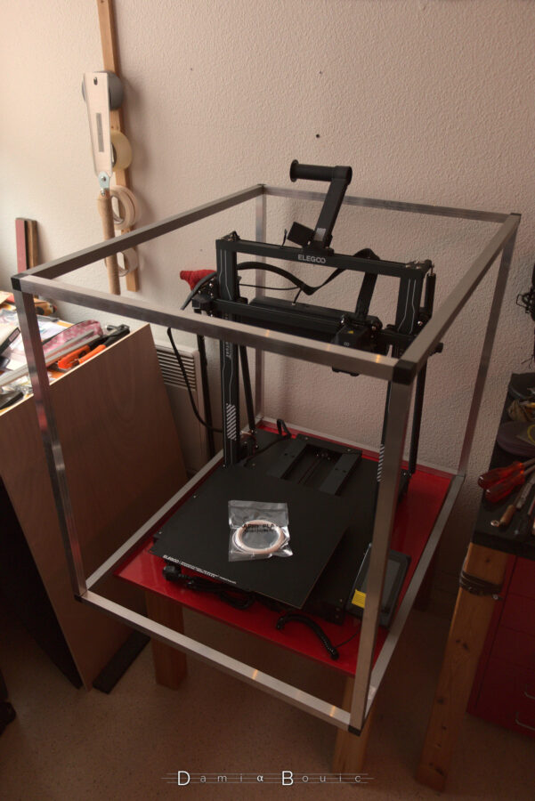 Le caisson dont on ne voit que les arrêtes en alu évidées. Dedans, l'imprimante 3D, qui aura été sortie de son carton et assemblée. Celle-ci ressemble à une balançoire dont le portique central et flanqué d'une bras sur courroie et la tête d'impression dessus. En bas, le lit d'impression, qui peut aller d'avant en arrière