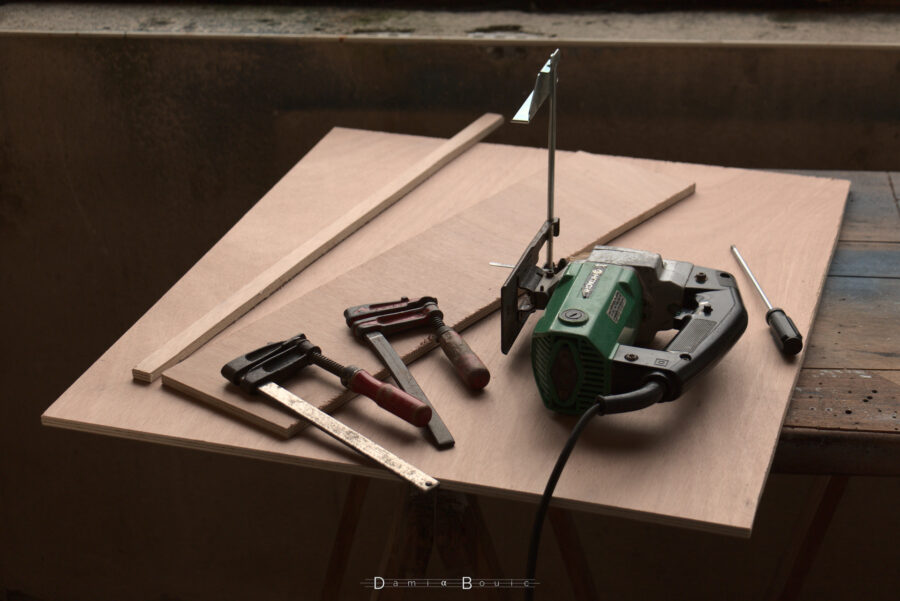 Retour balcon. Planche de contreplaqué, coupée aux bonnes dimensions. Deux chutes sont posées dessus, ainsi que deux serrre-joint et une scie sauteuse pourvue d'un guide en métal