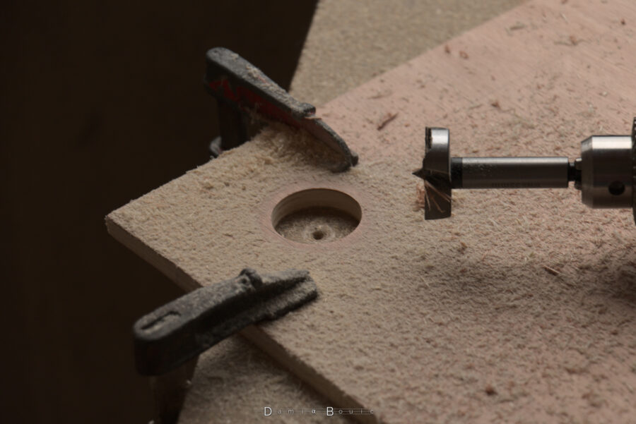 Détail sur un coin de la planche avec un large trou pratiqué par un forêt Forstner de grande dimension (35mm). Deux serre-joint maintiennent fermement la planche à l'établi, avec beaucoup de copeaux de bois partout.