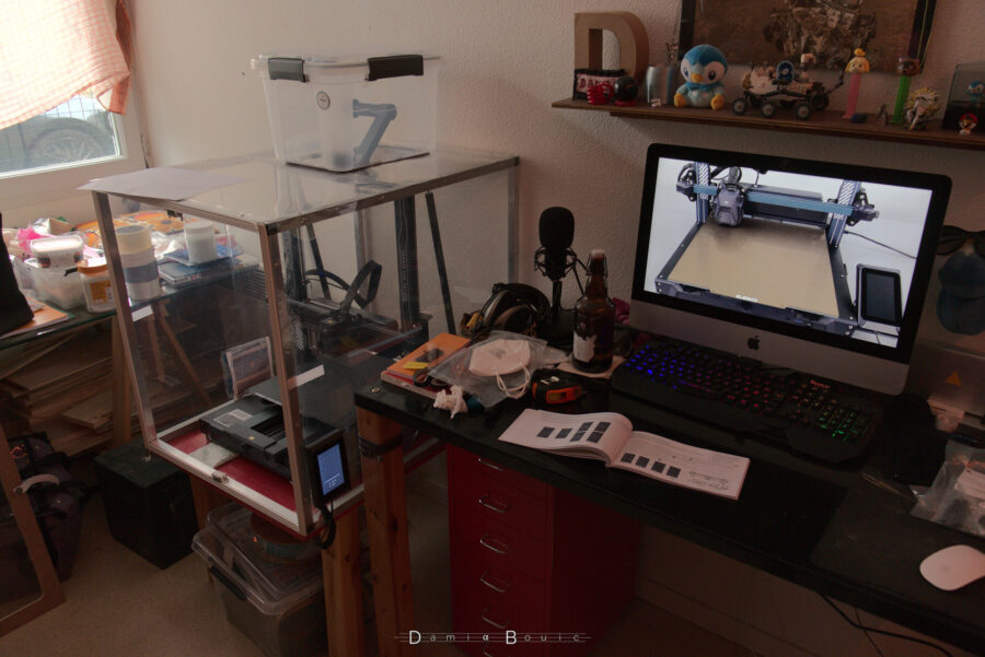 Vue générale de la mise en service de l'imprimante, un tutoriel vidéo sur l'écran de mon iMac, le livret d'utilisation sur le bureau, du bordel sur mon bureau aussi. A gauche, l'imposant caisson transparent de l'imprimante 3D, cette dernière étant affairée à calibrer des trucs.