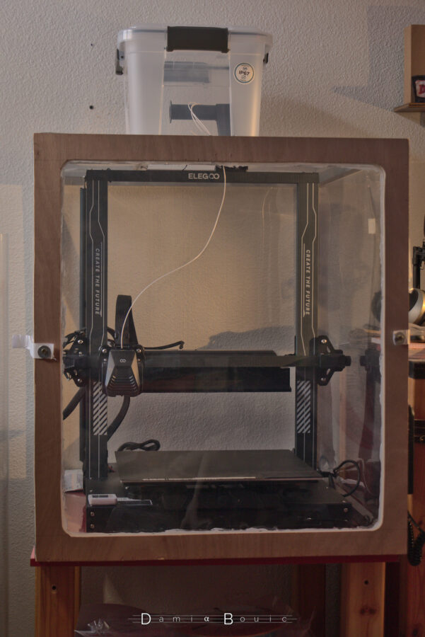 Vue finale, de face ou presque, sur l'imprimante 3D, dans son caisson, avec un filament qui alimente la buse d'impression.