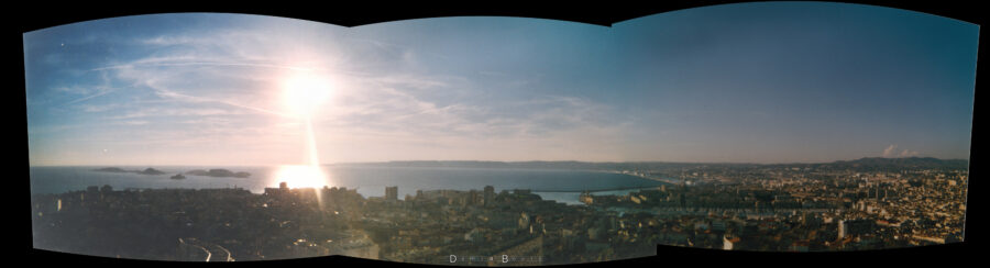 Panorama de 3 photos sur la ville de Marseille vue de Notre Dame de la Garde, avec le Soleil qui décline sur l'horizon, se reflétant fortement sur la mer. La ville est devant nous, en contrejours multiples de découpes de bâtiments, bordée par des collines plus loin et le trait de la Côte Bleue