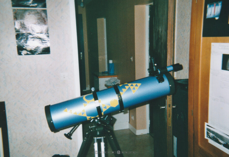 Mon télescope assemblé sur sa monture, dans ma chambre, dont on ne voit qu'une toute petite partie, devant la porte grande ouverte, dévoilant le couloir. Sur mon télescope des triangles jaunes autocollants colonisent le tube optique. Sur le mur à gauche, quelques photos de galaxies côtoient des photos de Saturne.