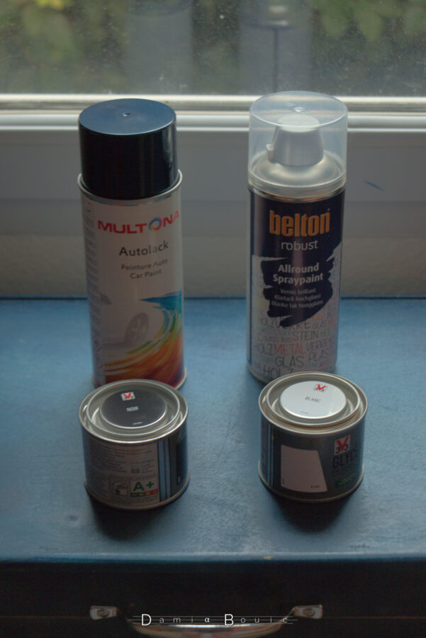 Deux bombes, l'une de peinture bleue métallisée dédiée pour l'étui et la couleur TARDIS, l'autre pour le dépôt d'un vernis transparent. Plus bas, deux petits pots de peinture laquée noir et blanc. Le tout est posé sur l'étui dont peut voir la poignée de transport chromée.