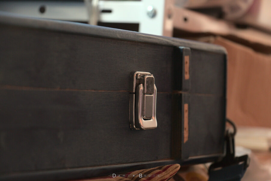 Loquet de fermeture brillant (ou chromé) vissé sur la fermeture. Il a l'allure d'un loquet qu'on trouverait sur un étui d'instrument de musique du commerce. Derrière, un peu dans le flou, un des anciens support des anciens loquets, qui ont été ôtés, laissant place à des petites zones de bois non peintes.