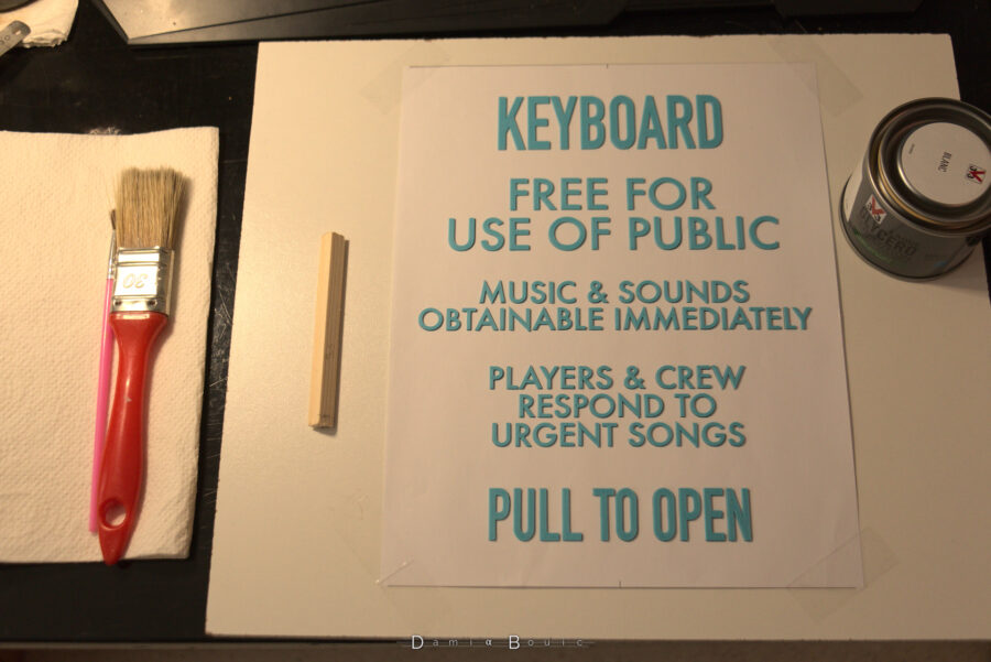 Préparation de texte en lettre plastique à être peint de couleur blanche, avec les pinceaux à gauche disposés sur du papier absorbant et à droite le pot de peinture.