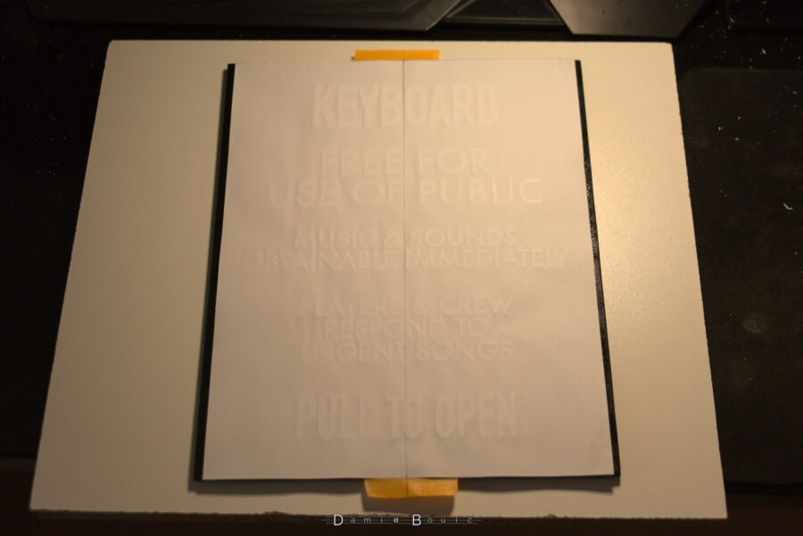 Début du collage définitif. La feuille est disposée à l'envers, les lettres posées sur la plaque en bois peinte en noir. La colle plus puissante permettra au texte d'adhérer plus à la plaque qu'à la feuille de papier.