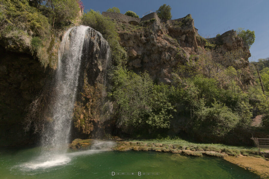 Une falaise rocheuse avec pas mal d'arbres, vue d'assez près, et une belle chute d'eau à gauche de l'image qui se déverse dans un plan d'eau, sorte de mare, presque circulaire