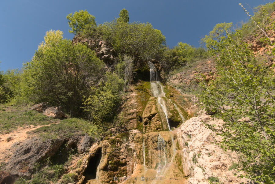 Tableau contrasté de roches exposées, de roches moussues, avec des filets d'eau qui y coulent, en cascade discrète. Des arbres accrochés aux rochers entourent le tout.