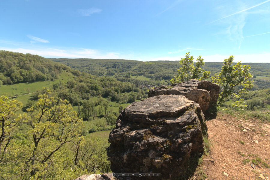 Deux gros rochers anguleux, sombres et recouverts de mousses et lichens servent d'ancre au premier plan, l'arrière plan étant constitué d'une belle vue sur la vallée et les collines forestières, sous un Soleil écrasant et un ciel bleu insolent barré ci et là de trainées de condensations
