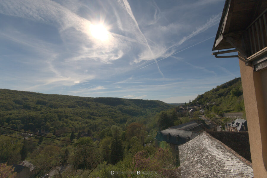 Vue à contrejour sur la vallée boisée, au dessus de quelques toits en ardoise, qui reflètent fortement la lumière solaire. De plus en plus de cirrus occupent le ciel.