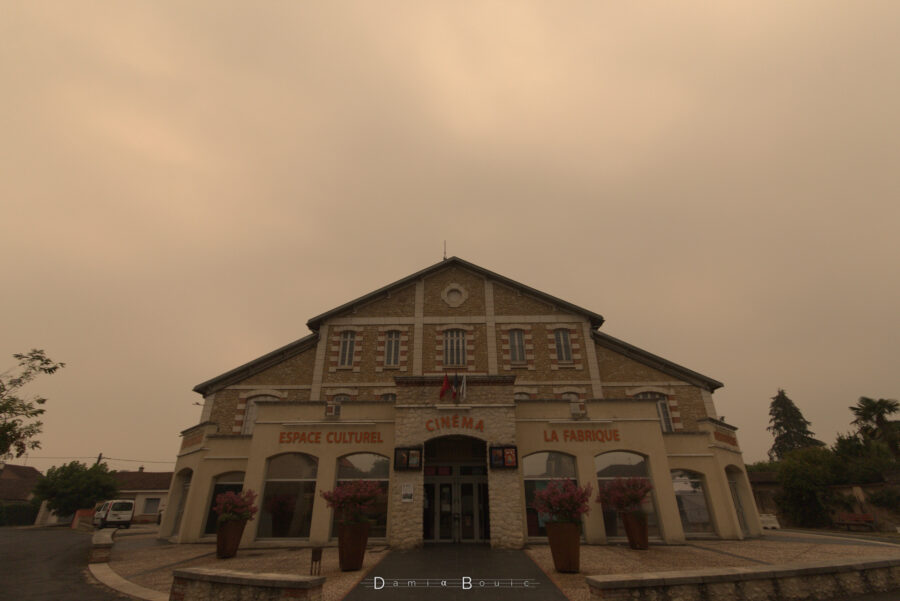 Le bâtiment de la Fabrique à Saint-Astier, aux formes très évocatrices d'une ancienne manufacture, toiture imposante et triangulaire vue de face, larges fenêtres, notamment au rez-de-chaussée, avec les inscriptions Espace Culture, Cinéma, La Fabrique. Tout ceci sous un ciel nuageux jaunâtre très inhabituel