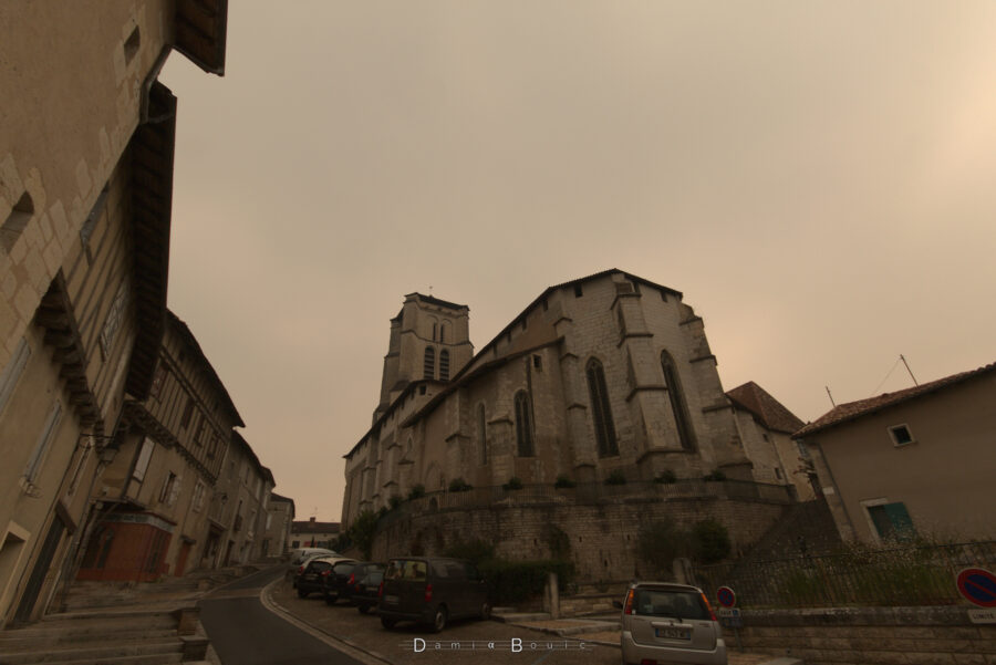 Rue en contrebas de l'imposante église de Saint-Astier, sorte de gros bâtiment en pierre d'allure médiévale avec quelques mises à jour plus modernes de l'époque gothique. La rue à gauche monte devant des façades anciennes. Le tout sous un ciel chargé de fumées de feux forestiers