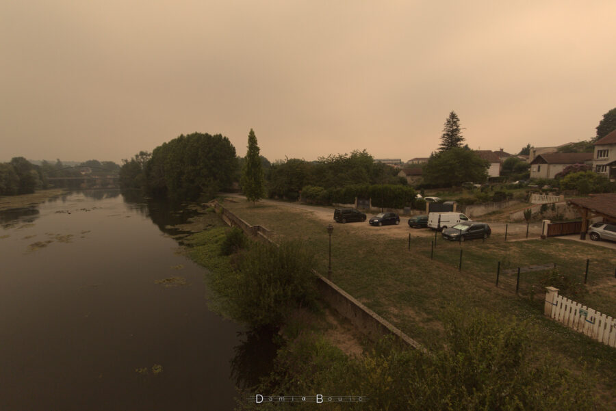 Vue presque identique, mais plus centrée sur la rive droite, avec des pelouses rases et quelques voitures garées devant des portails d'habitations.