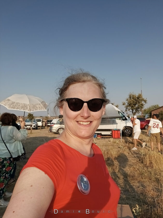 Autoportrait photo de l'autrice de ce blog, à savoir moi-même, tout sourire, face au Soleil, lunettes de Soleil, tee-shirt rouge et badge de mission fièrement arboré. En arrière-plan, l'agitation de ce vrai mini village astro