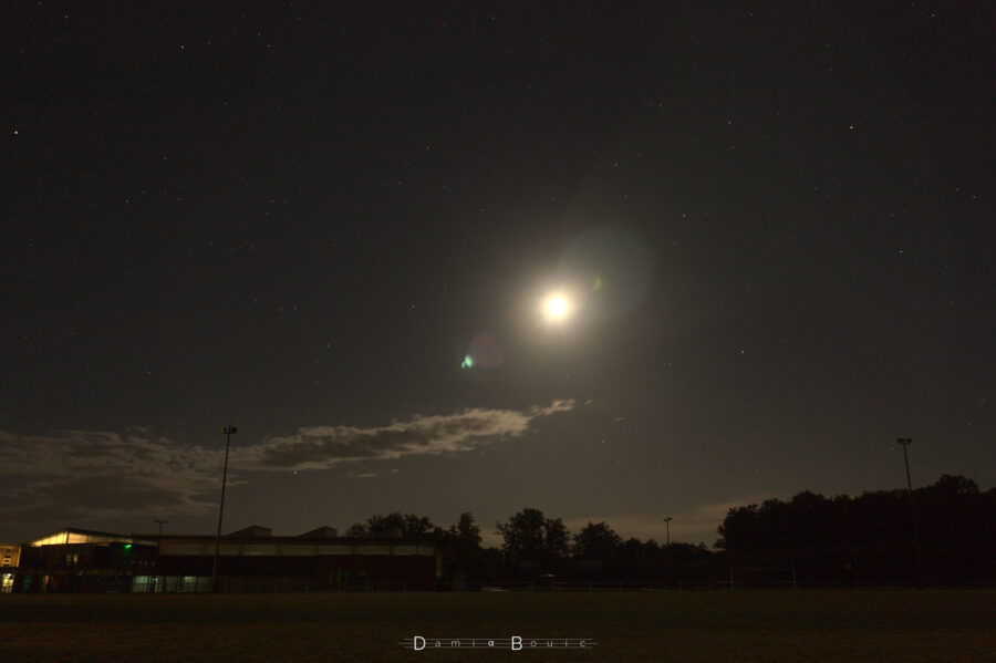 Photo à grand angle au bord d'un terrain de foot d'établissement scolaire, quelques bâtiments et une poignée d'arbres forment l'horizon. Dans le ciel étoilé au dessus trône la Lune, et un banc de nuages près de l'horizon