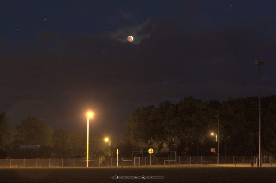 Zoom juste avant que la Lune ne soit envahie de nuages. C'est le maximum, on parvient à voir le disque rouge de la Lune et son tout petit bout encore illuminé. Des lampadaires se sont allumés en dessous et éclairent une route vide.