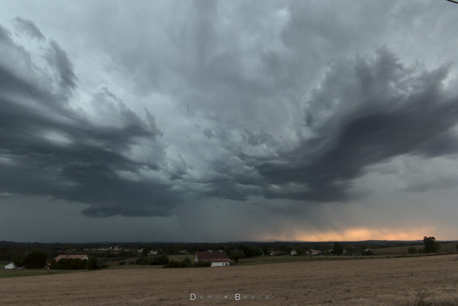 En ultra grand angle, ciel orageux et chaotique de nuages menaçants. L'horizon est sombre et des rideaux de pluie sont visibles, passant devant les Pyrénées, avec les couleurs du couchant au travers 