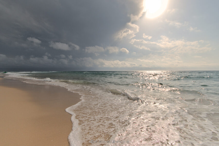 Bord de mer, avec l'eau qui s'est abattue sur le sable luisant et étincelant, laissant beaucoup d'écume. Le tout sous un Soleil présent mais qui commence à être masqué par l'enclume d'un nuage d'orages qui nait bien plus loin, à gauche au delà de l'horizon