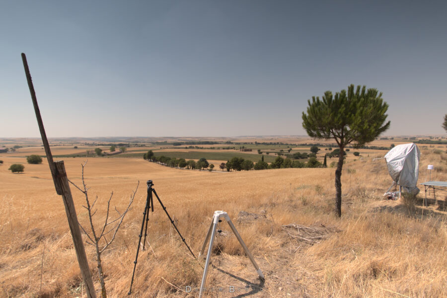 Photo de mon futur poste de travail pour l'éclipse. Mon trépied astro et mon trépied photo sont posés côte-à-côte, et la vue se déploie sur le plateau semi-aride espagnol, sous un Soleil de plomb. A droite, un tout petit pin et un instrument placé sous une bâche de protection
