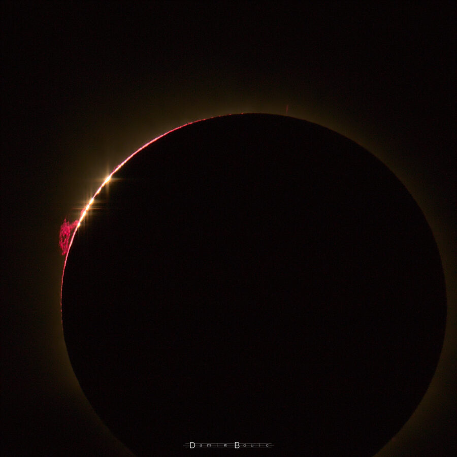 D'ultimes grains de lumière se manifestent sur le bord lunaire. Une protubérance rouge vif est visible à gauche.