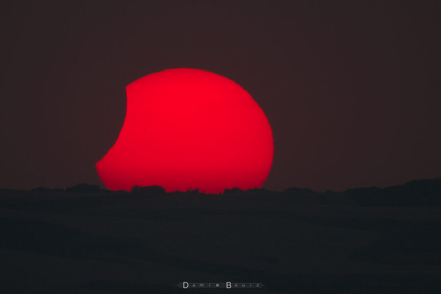 Coucher de Soleil éclipsé. Les bords du Soleil sont agités, il ne reste plus qu'un morceau de lune à gauche sur l'astre. Le profil de l'horizon se dessine sur le bas de l'astre du jour